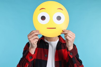 Man covering face with surprised emoticon on light blue background