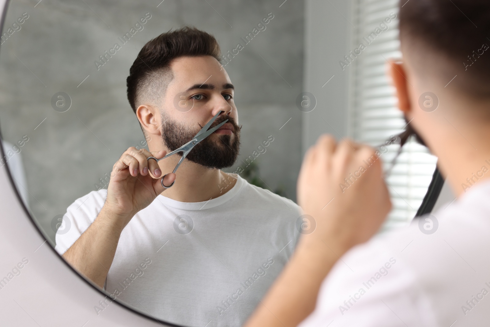 Photo of Handsome young man trimming mustache with scissors near mirror in bathroom