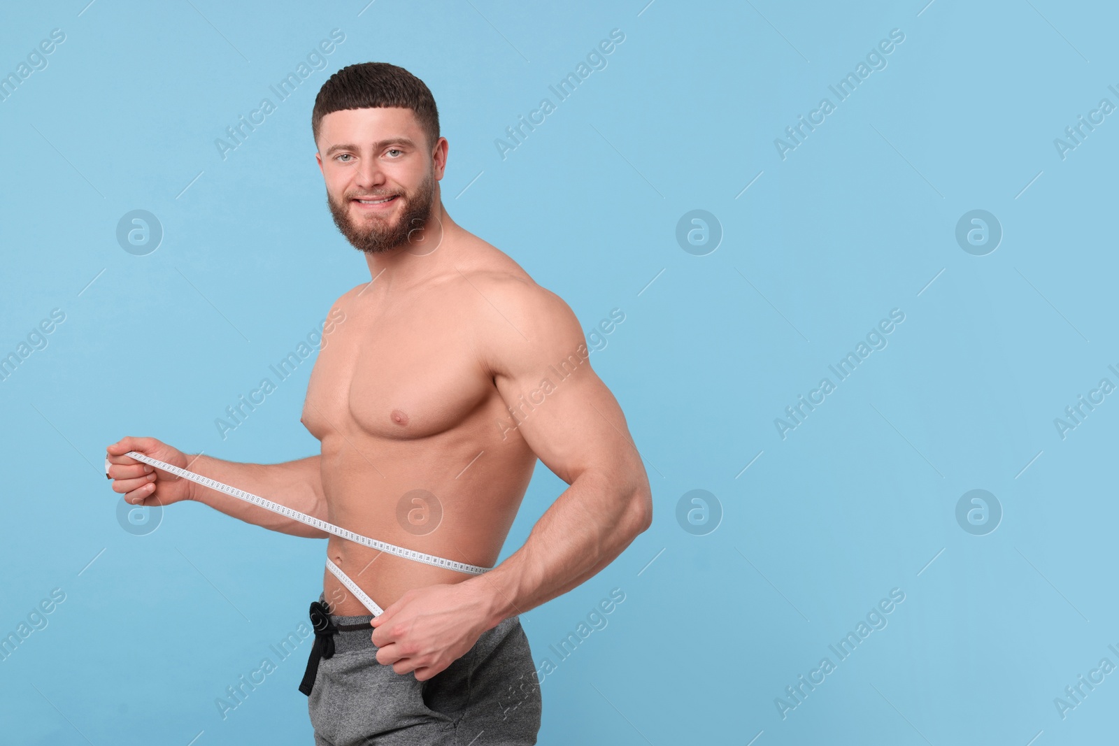 Photo of Portrait of happy athletic man measuring waist with tape on light blue background, space for text. Weight loss concept