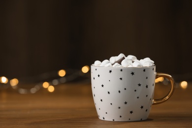 Cup of hot drink with marshmallows and chocolate on wooden table against blurred lights. Space for text