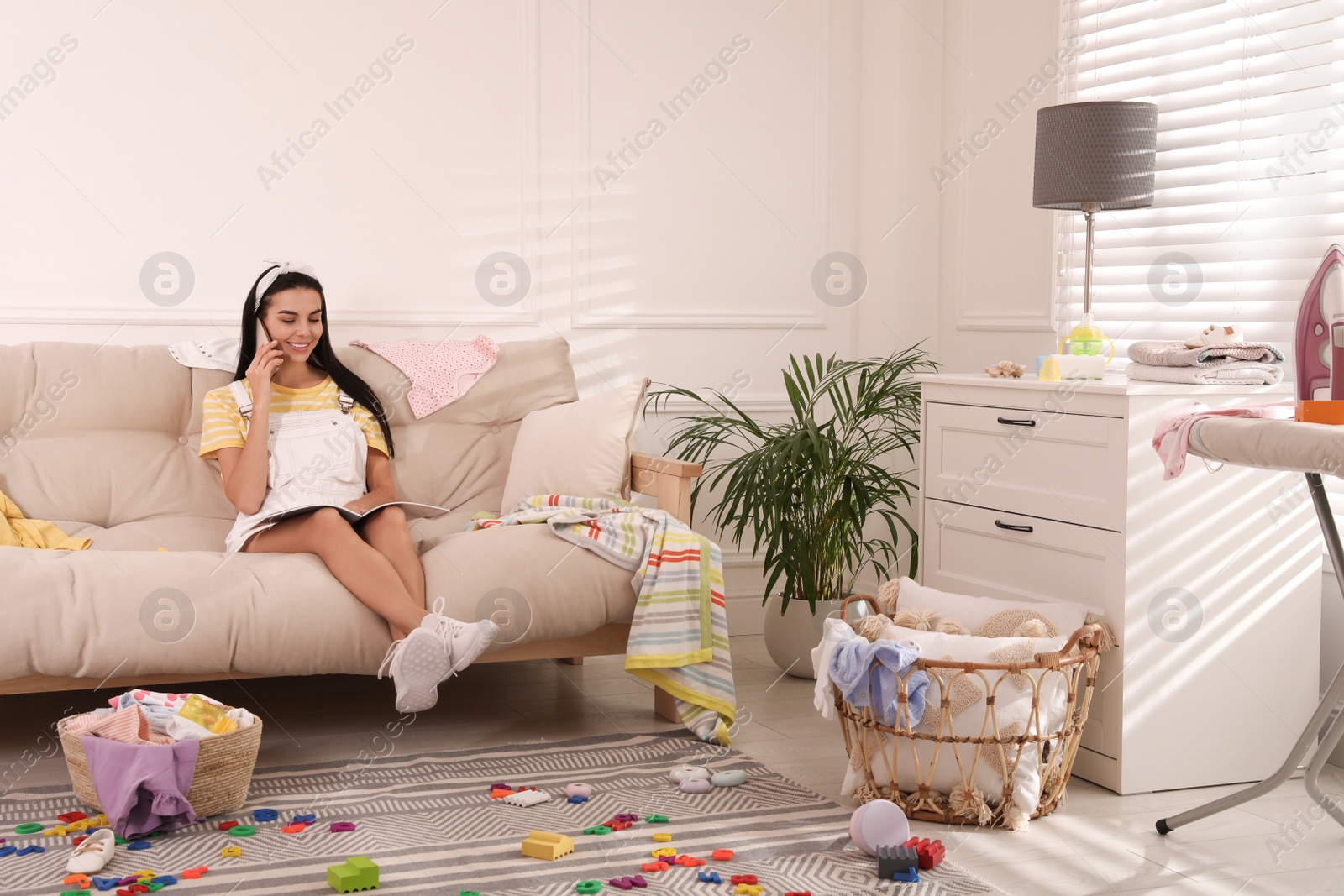 Photo of Young mother with magazine talking by mobile phone on sofa in messy living room