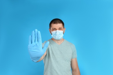 Photo of Man in protective mask showing stop gesture on light blue background. Prevent spreading of COVID‑19