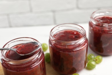 Jars of delicious gooseberry jam and fresh berries on white table, closeup