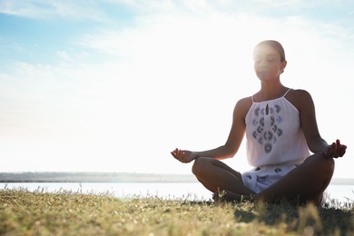 Young woman meditating near river at sunset, space for text. Nature healing power