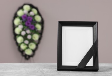 Photo of Photo frame with black ribbon on table and wreath of plastic flowers near grey wall, space for text. Funeral attributes