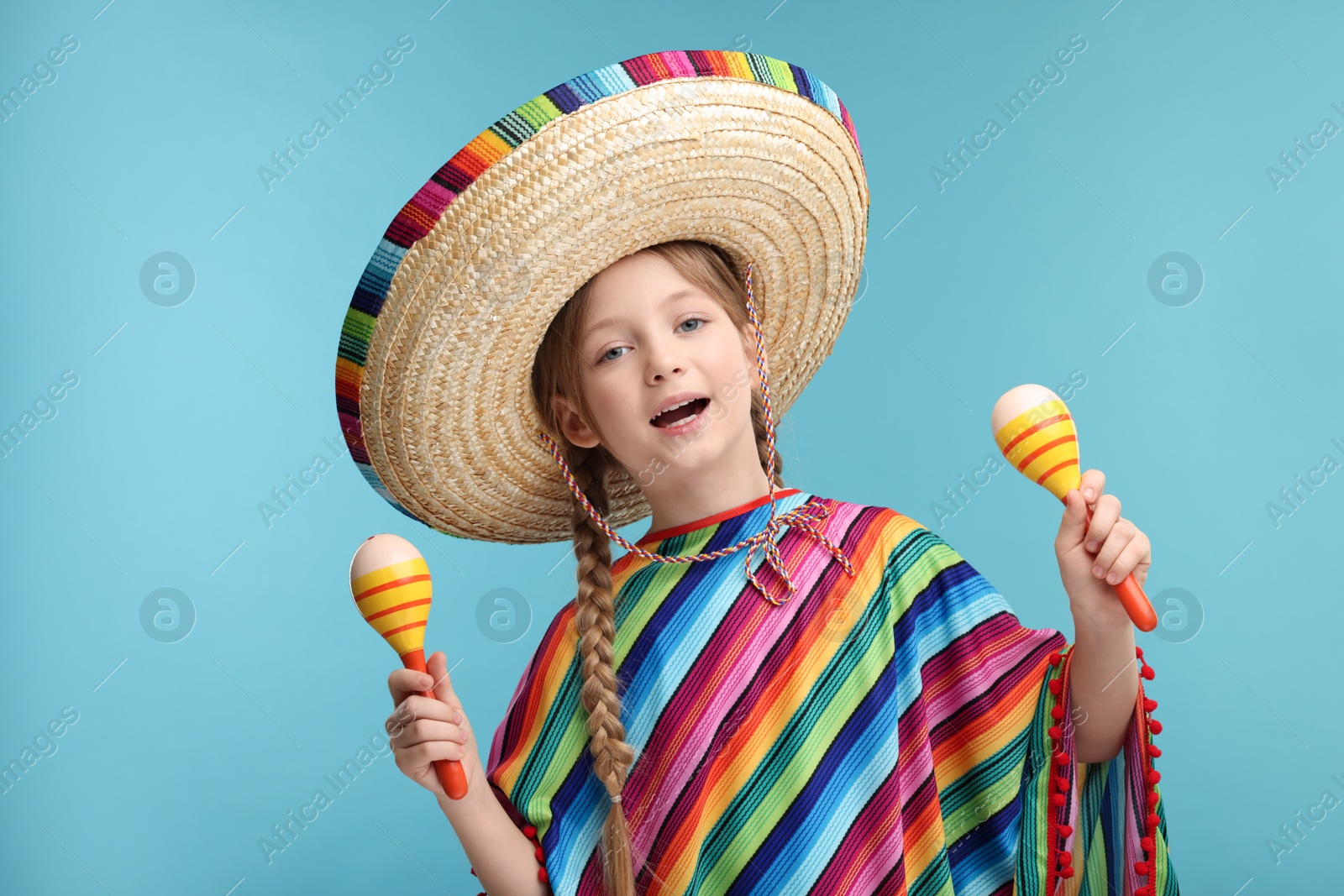 Photo of Cute girl in Mexican sombrero hat and poncho dancing with maracas on light blue background