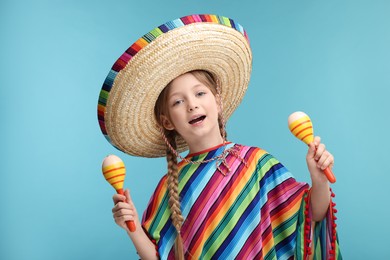 Cute girl in Mexican sombrero hat and poncho dancing with maracas on light blue background