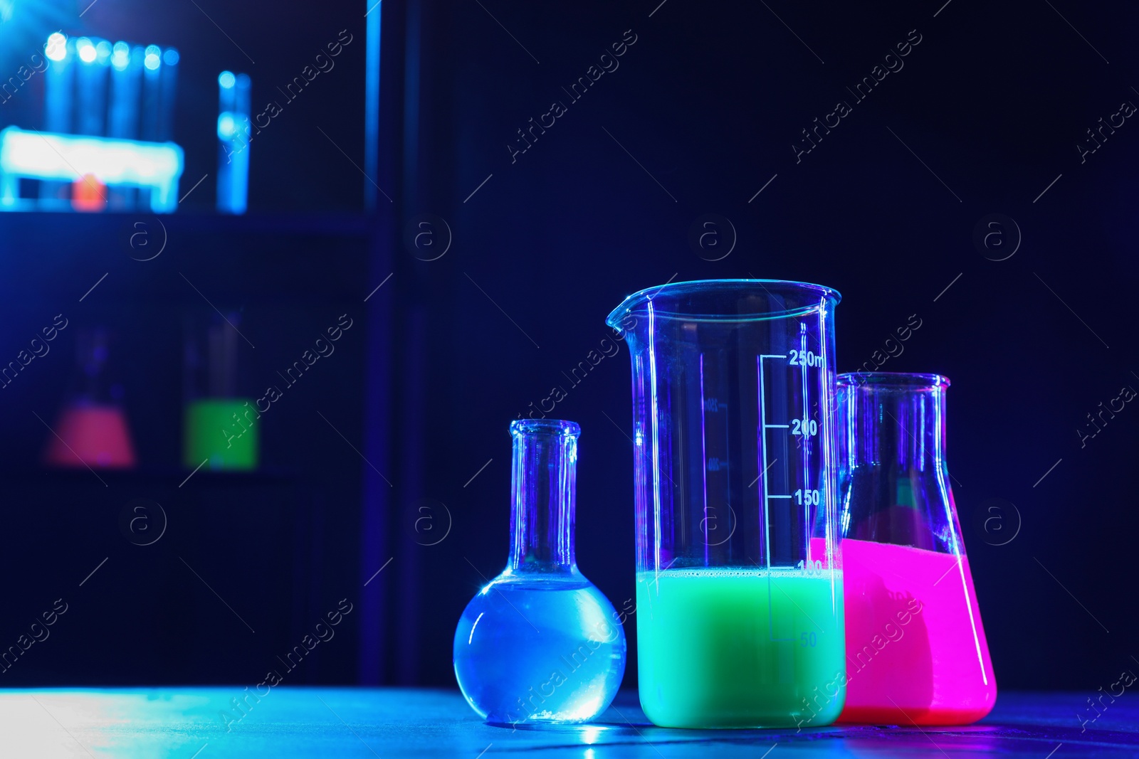 Photo of Laboratory glassware with luminous liquids on table, space for text