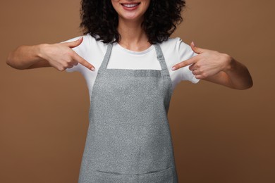 Woman pointing at kitchen apron on brown background, closeup. Mockup for design