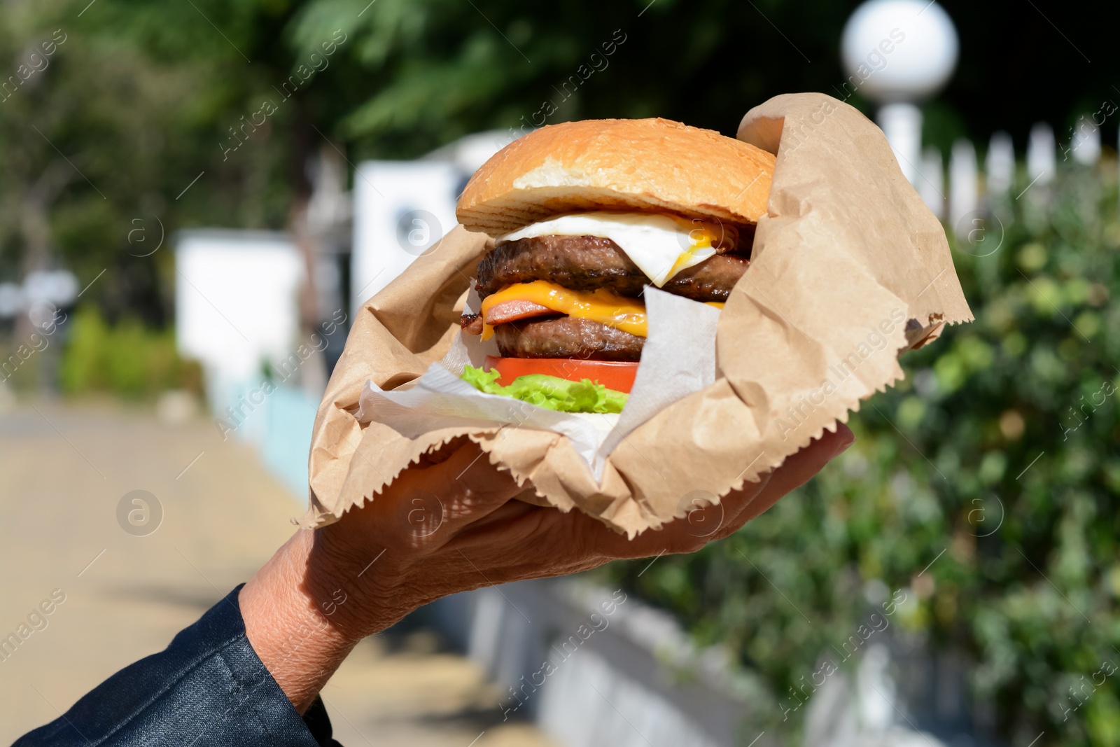 Photo of Woman holding delicious burger in paper wrap on city street, closeup