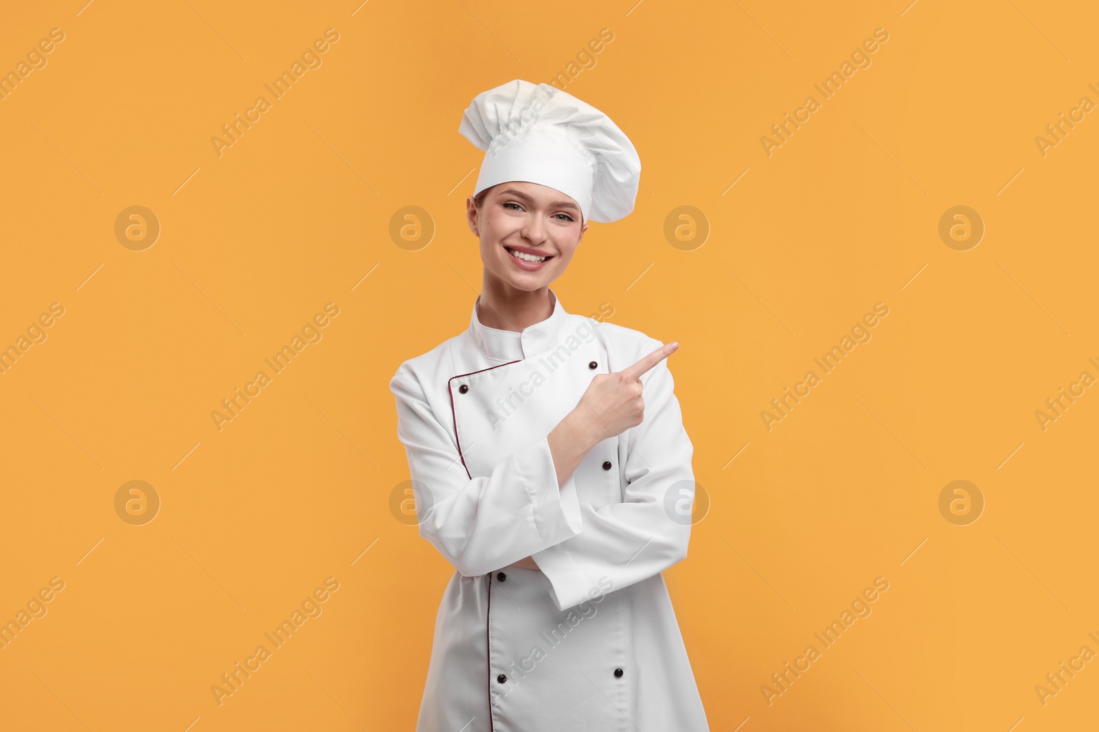 Photo of Happy woman chef in uniform pointing at something on orange background, space for text