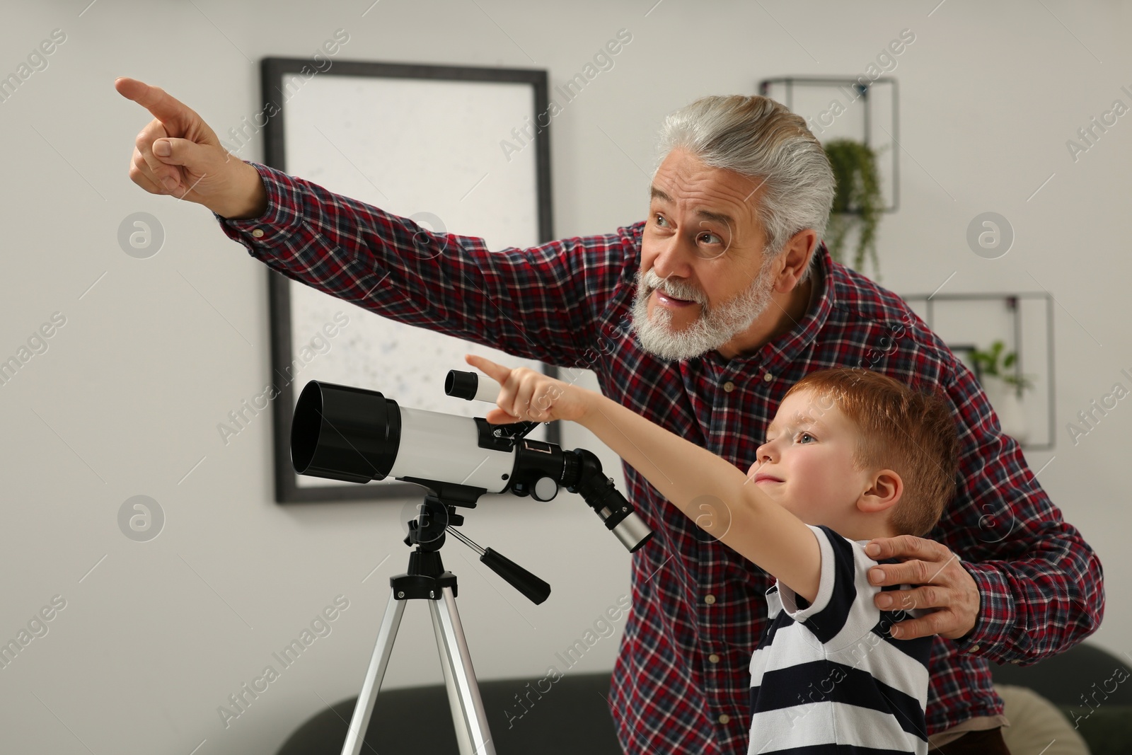 Photo of Little boy with his grandfather pointing at something near telescope in room