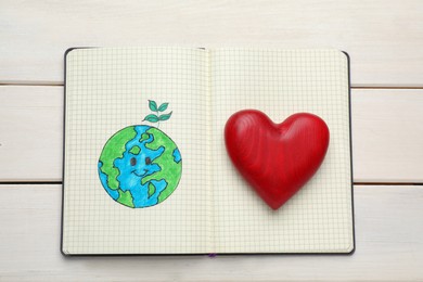 Photo of Happy Earth Day. Notebook with drawing of planet and decorative heart on white wooden table, top view