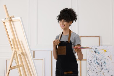 Photo of Young woman holding brush and artist`s palette near easel with canvas