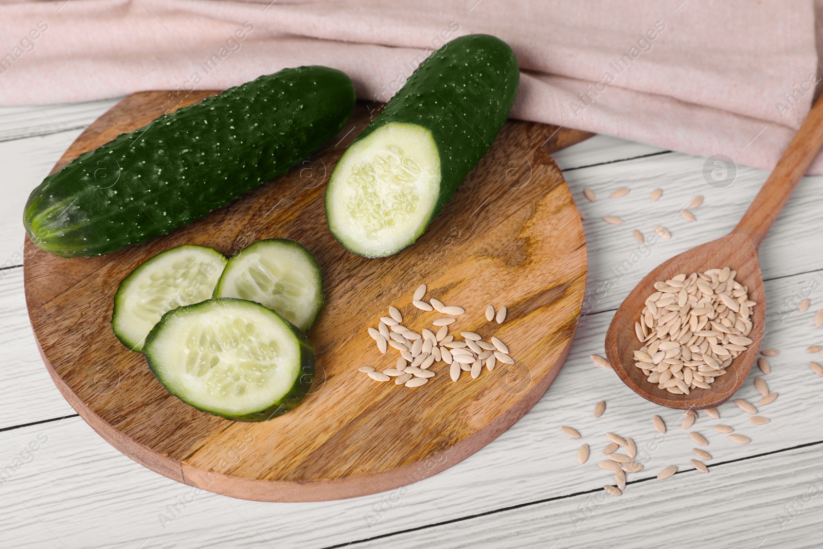 Photo of Spoon of vegetable seeds and fresh cucumbers on white wooden table, above view