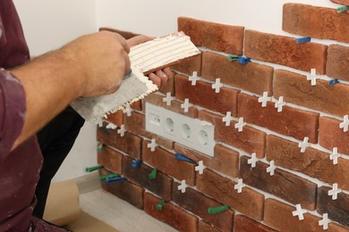 Photo of Professional builder applying cement onto decorative brick near wall, closeup. Tiles installation process
