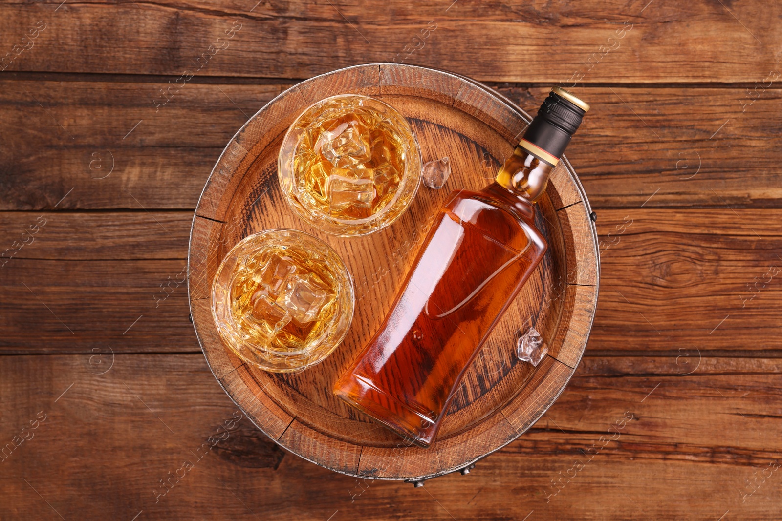 Photo of Whiskey with ice cubes in glasses, bottle and barrel on wooden table, top view