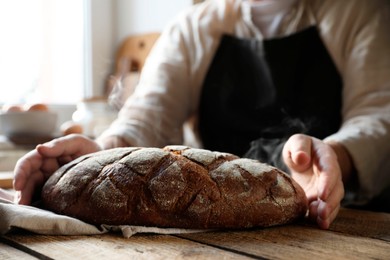 Photo of Man holding loaf of fresh bread at wooden table indoors, closeup