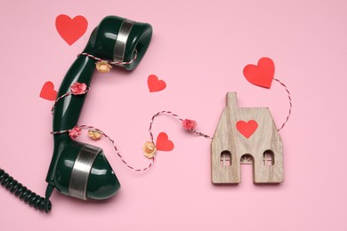 Photo of Decorative cord and paper hearts between telephone receiver and wooden house model on pink background symbolizing connection in long-distance relationship, flat lay
