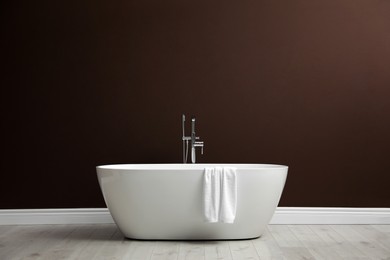 Modern ceramic bathtub with towel near brown wall indoors