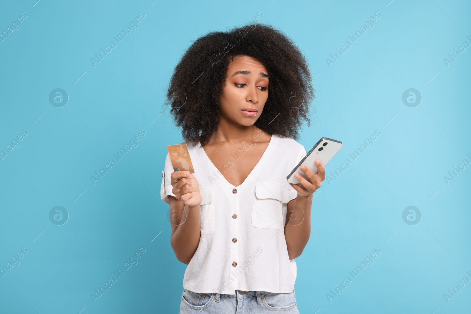 Photo of Confused woman with credit card and smartphone on light blue background. Debt problem
