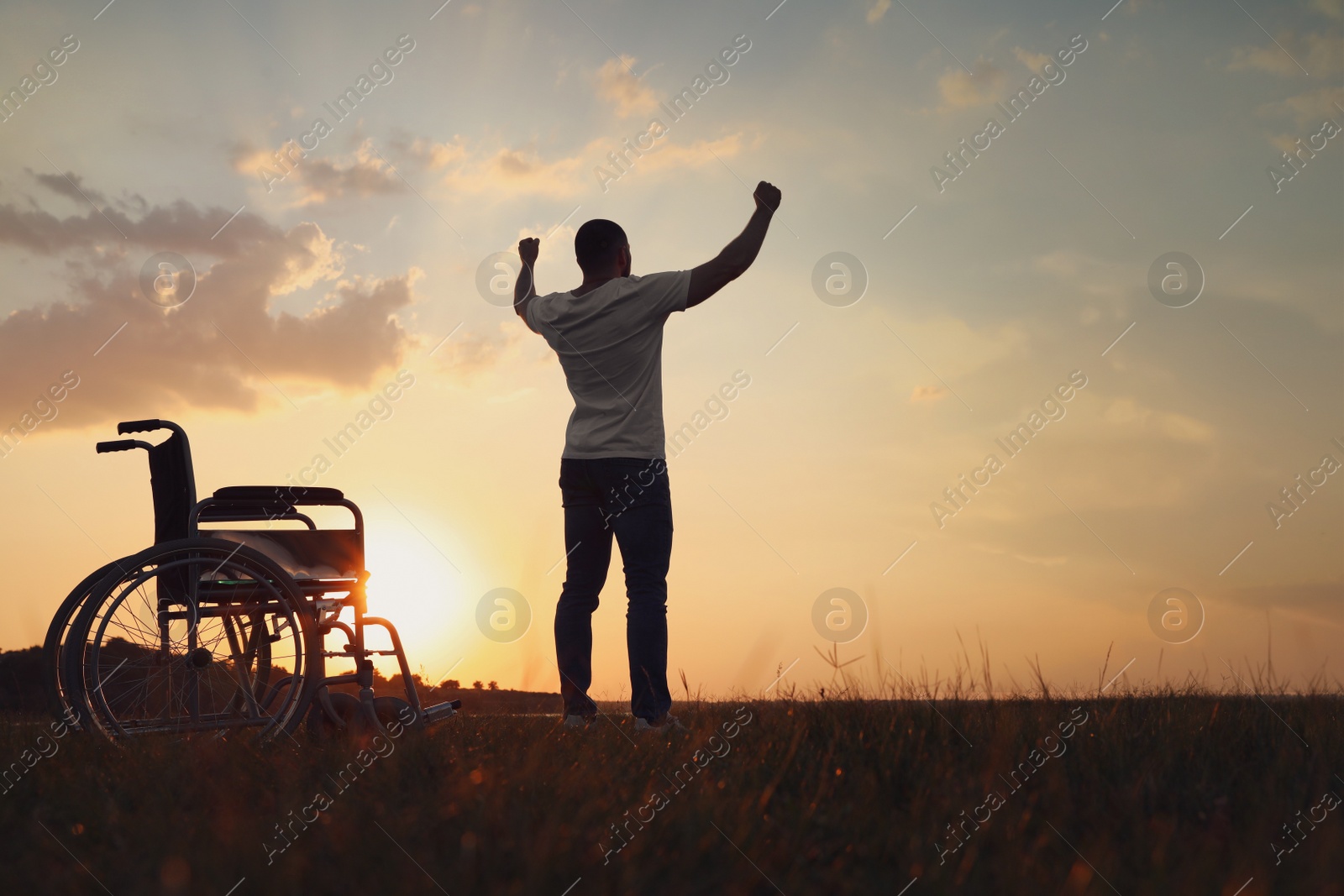 Photo of Man raising hands up to sky near wheelchair at sunset, back view. Healing miracle