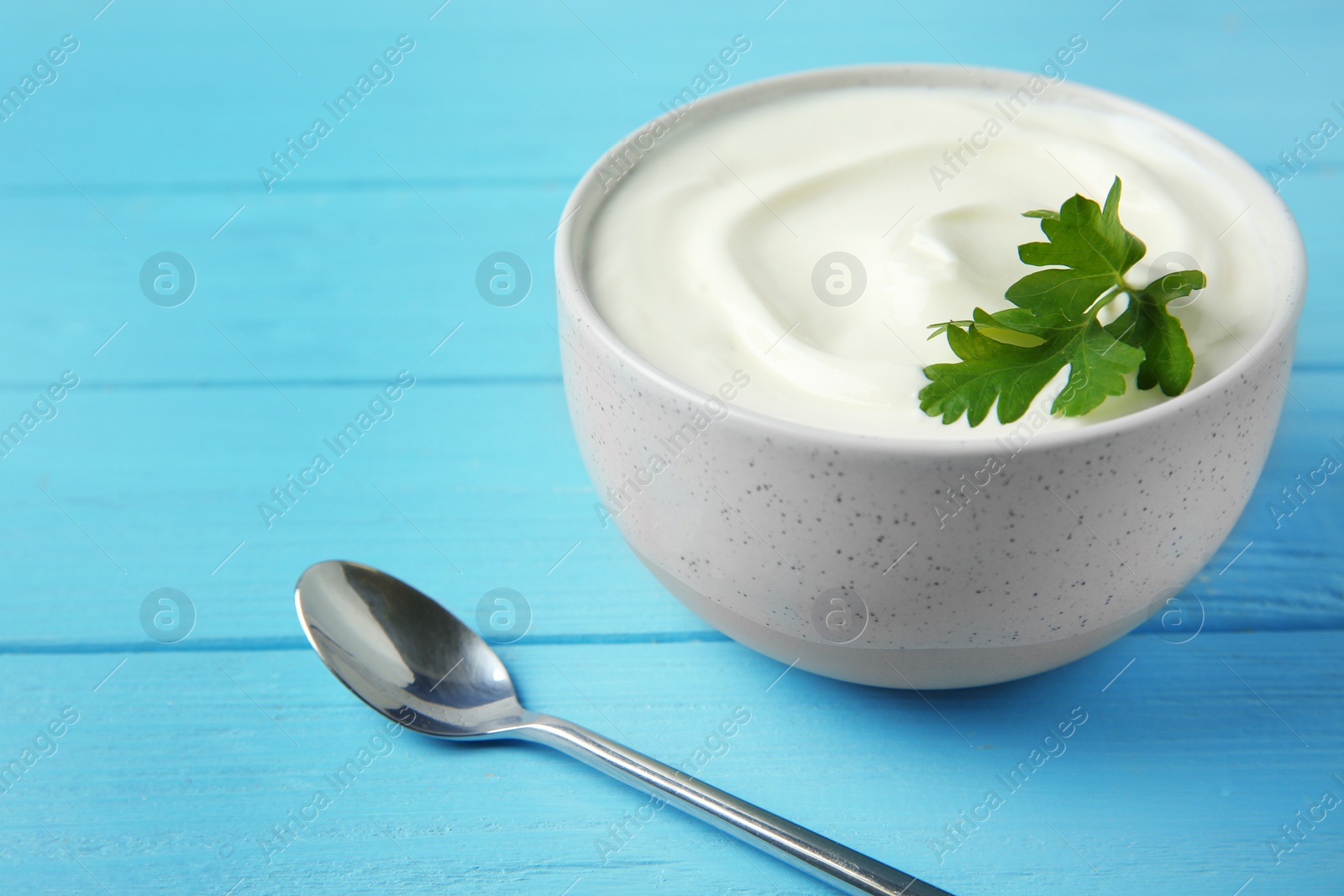 Photo of Bowl of fresh sour cream with parsley and spoon on light blue wooden table, space for text