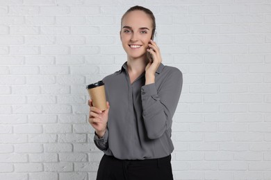 Photo of Happy young secretary with paper cup of drink talking on smartphone near white brick wall