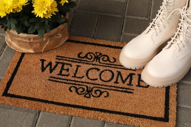 Photo of Door mat with word Welcome, stylish boots and beautiful flowers on floor