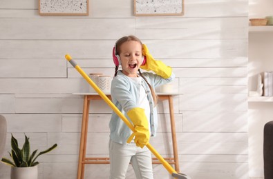 Photo of Cute little girl in headphones with mop singing while cleaning at home