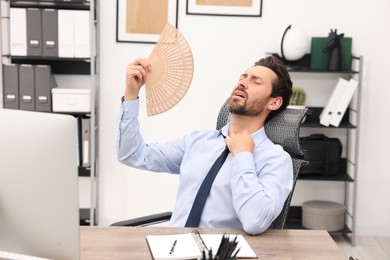 Photo of Bearded businessman waving hand fan to cool himself at table in office