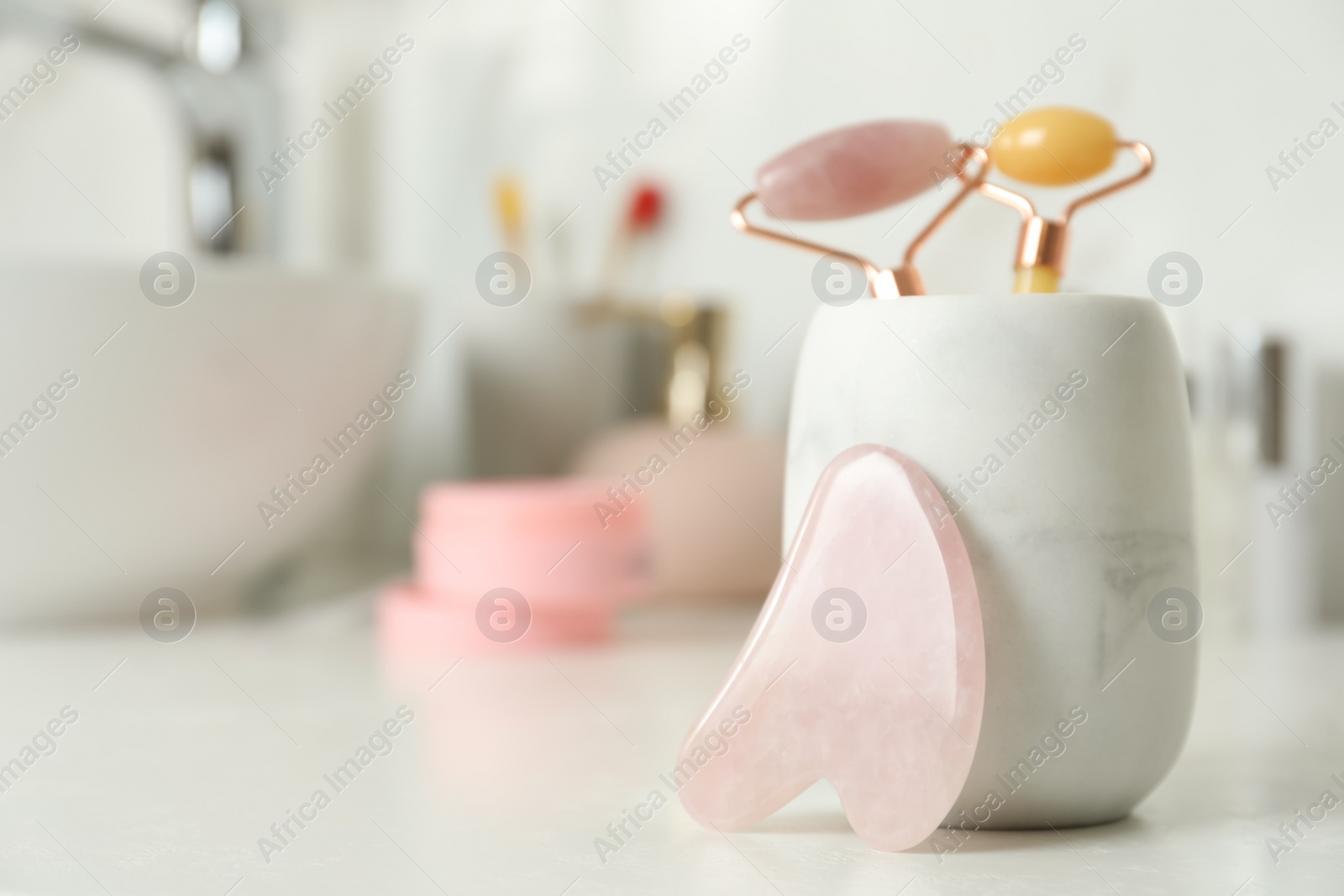 Photo of Rose quartz gua sha tool near holder with natural face rollers on white countertop in bathroom. Space for text