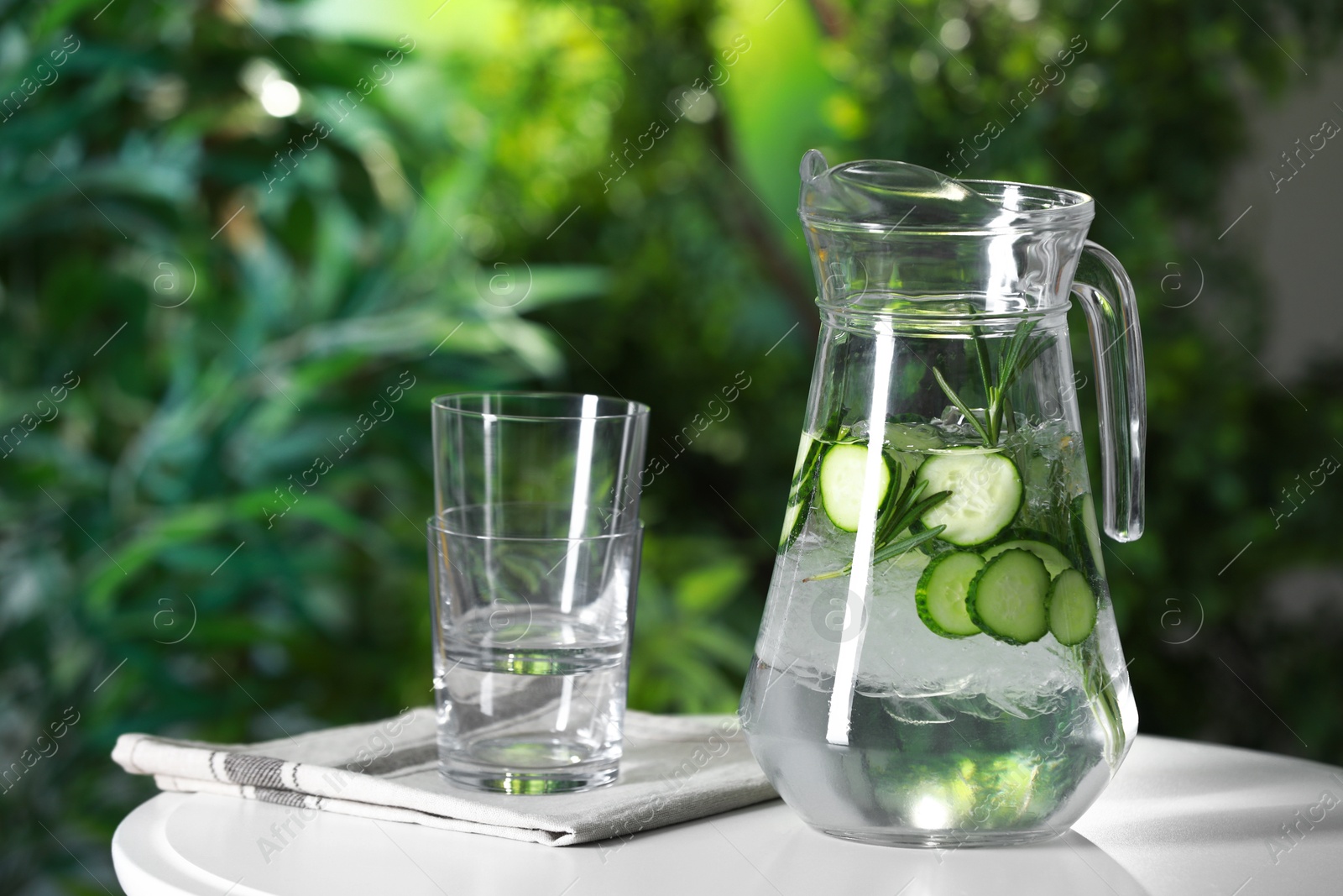 Photo of Refreshing cucumber water with rosemary in jug and glasses on white table against blurred green background