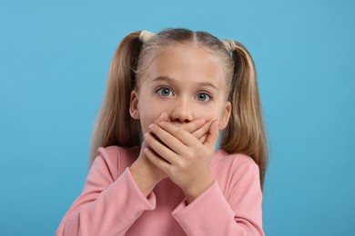 Photo of Embarrassed little girl covering her mouth with hands on light blue background