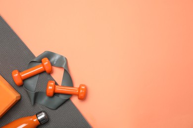 Dumbbells, thermo bottle, fitness elastic band, yoga block and mat on coral background, flat lay. Space for text