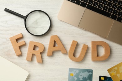 Word Fraud made of wooden letters, credit cards, laptop and magnifying glass on white table, flat lay