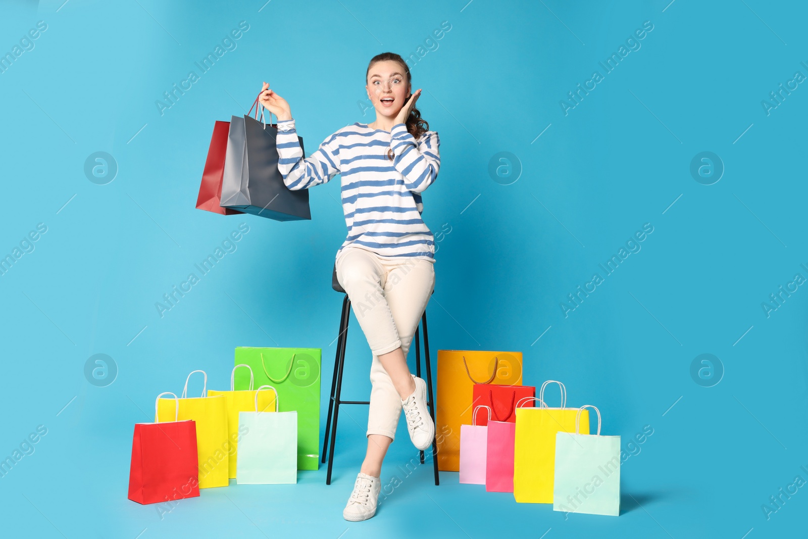 Photo of Excited woman holding colorful shopping bags on stool against light blue background