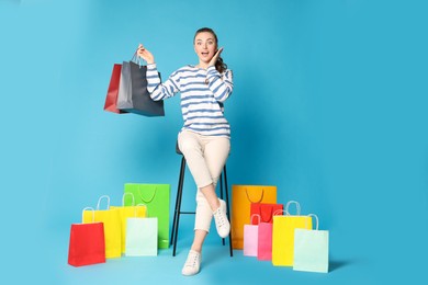 Excited woman holding colorful shopping bags on stool against light blue background