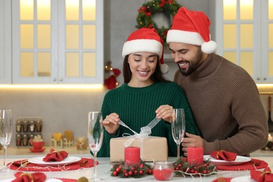 Happy young woman in Santa hat opening Christmas gift from her boyfriend at table in kitchen