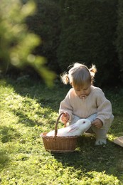 Photo of Cute little girl with adorable rabbit in wicker basket outdoors on sunny day