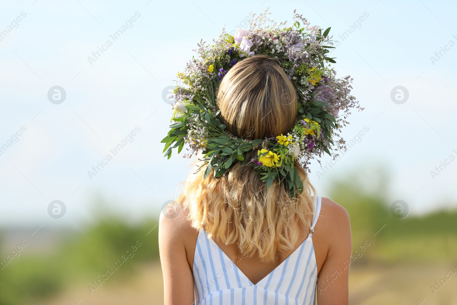 Photo of Young woman wearing wreath made of beautiful flowers outdoors on sunny day, back view