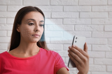 Image of Young woman unlocking smartphone with facial scanner near white brick wall. Biometric verification