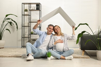 Photo of Young family housing concept. Pregnant woman with her husband sitting under cardboard roof on floor at home