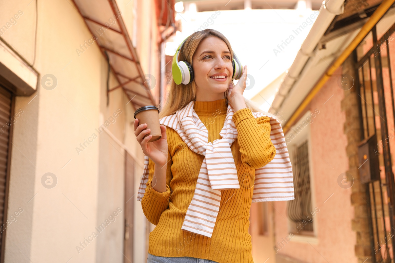 Photo of Happy young woman with coffee and headphones listening to music on city street