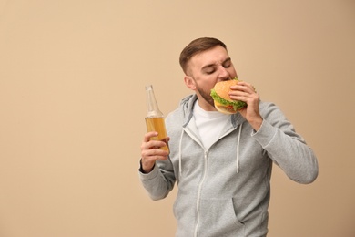 Young man with beer eating tasty burger on color background. Space for text