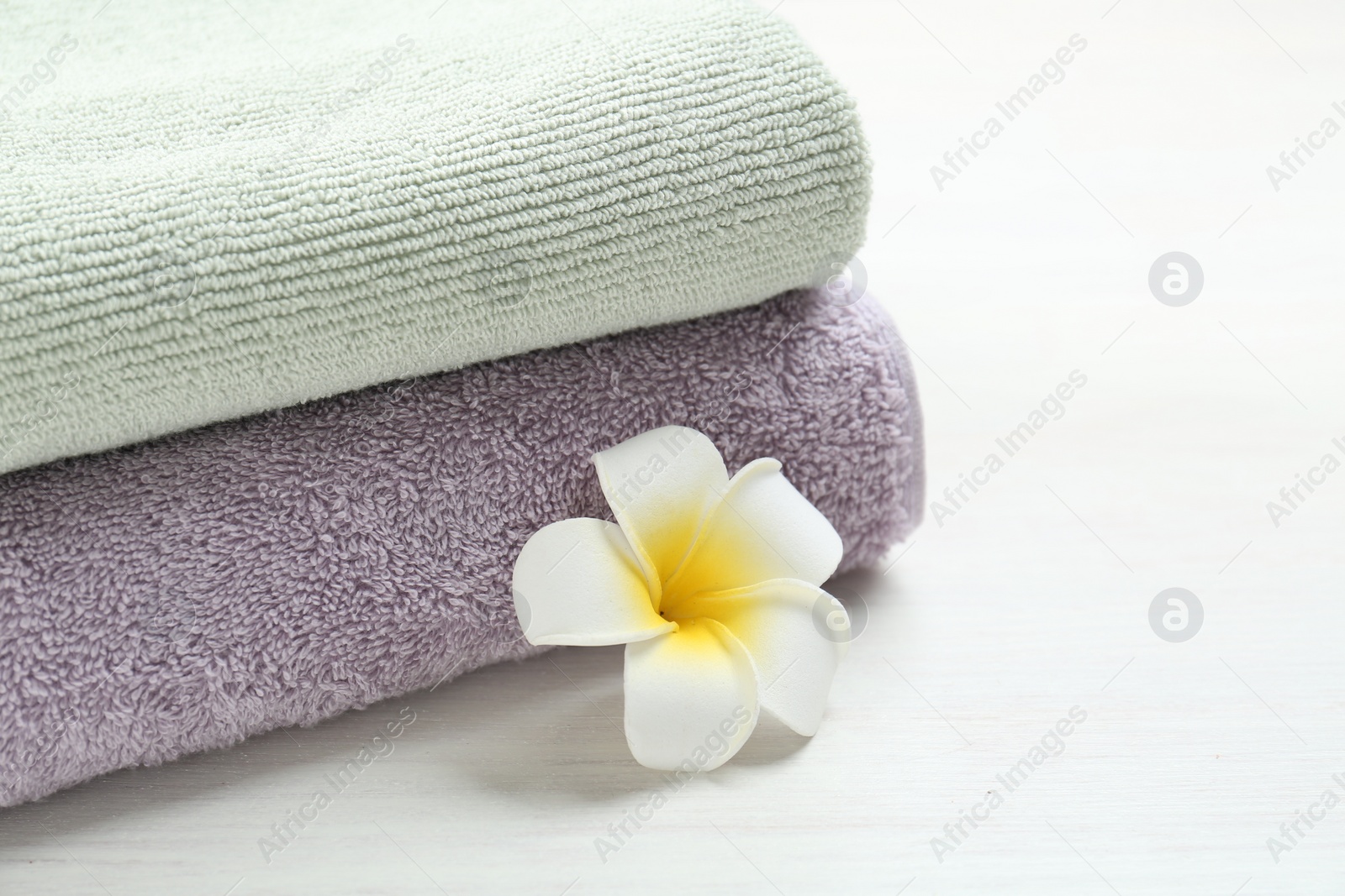 Photo of Different folded towels and plumeria flower on white wooden table, closeup. Space for text