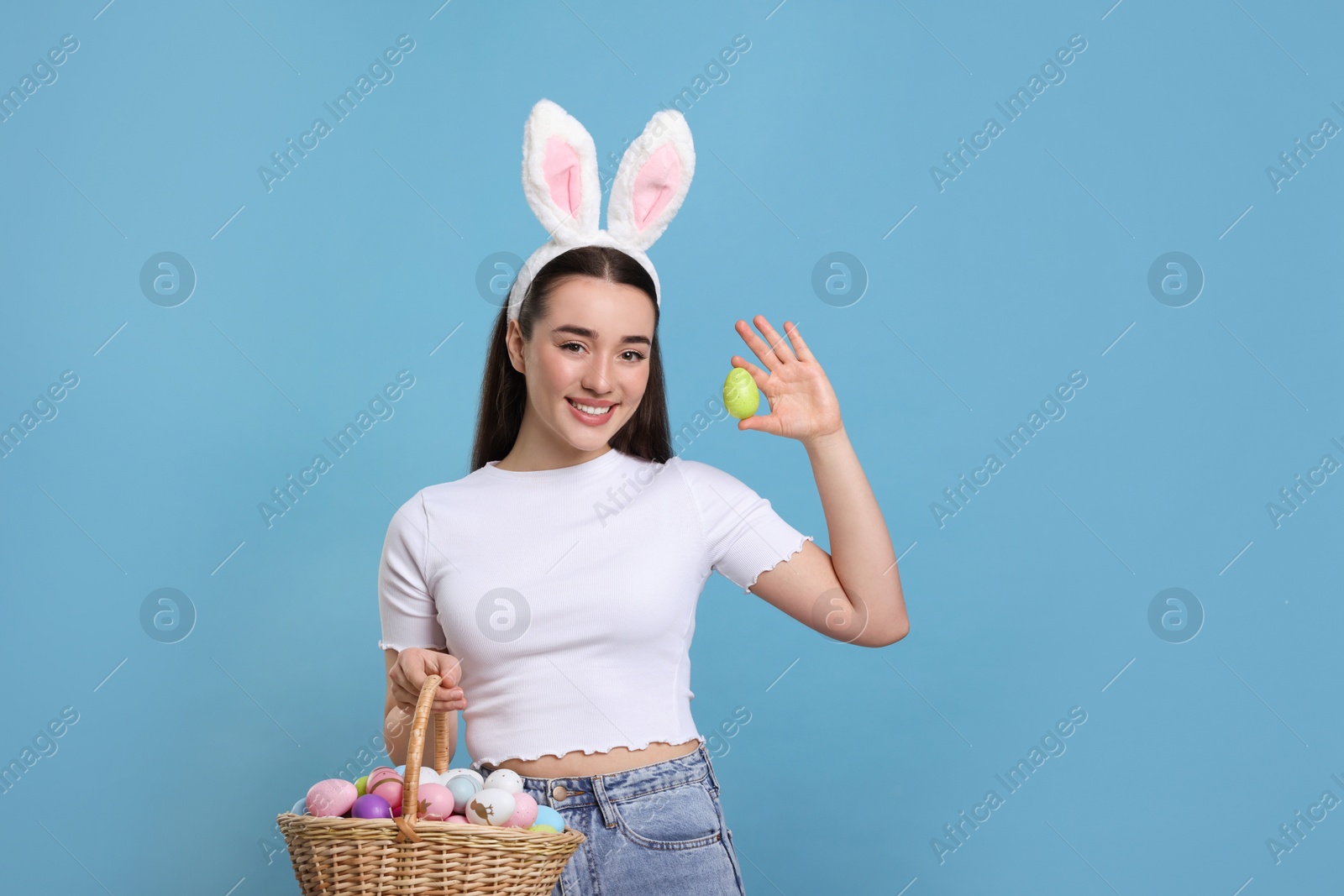 Photo of Happy woman in bunny ears headband holding wicker basket of painted Easter eggs on turquoise background. Space for text