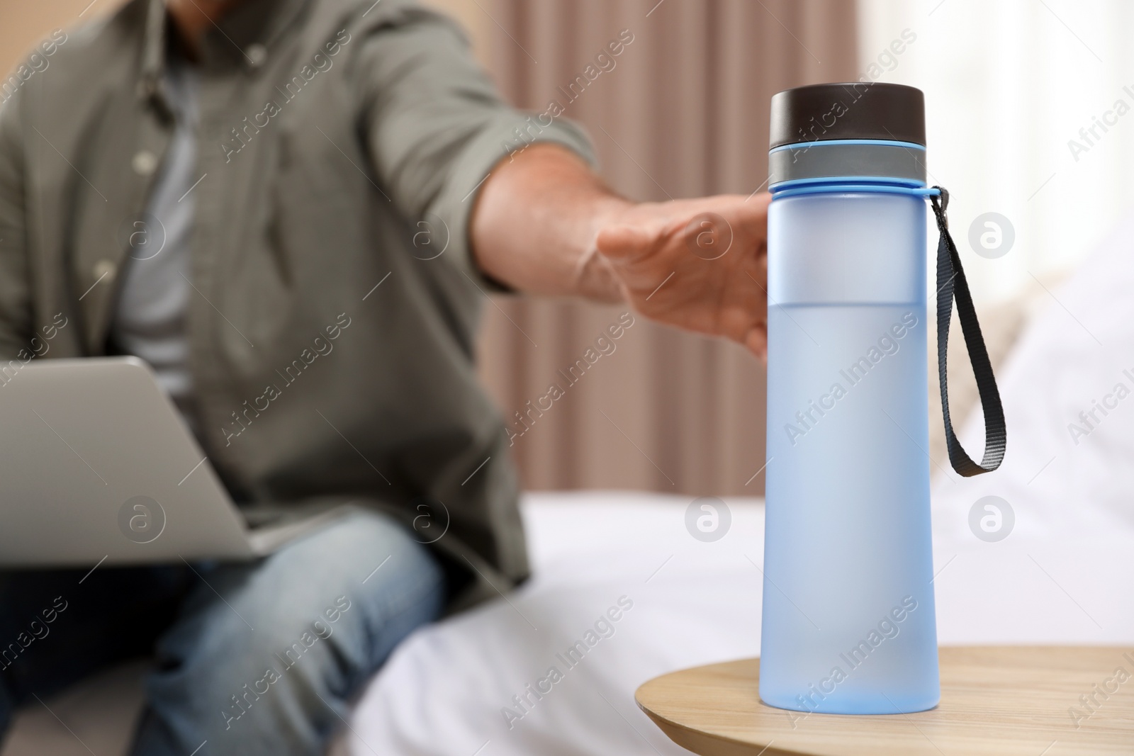 Photo of Man taking transparent plastic bottle of water while working on laptop indoors, closeup