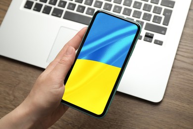 Image of Woman holding smartphone with Ukrainian national flag on screen at wooden table, closeup