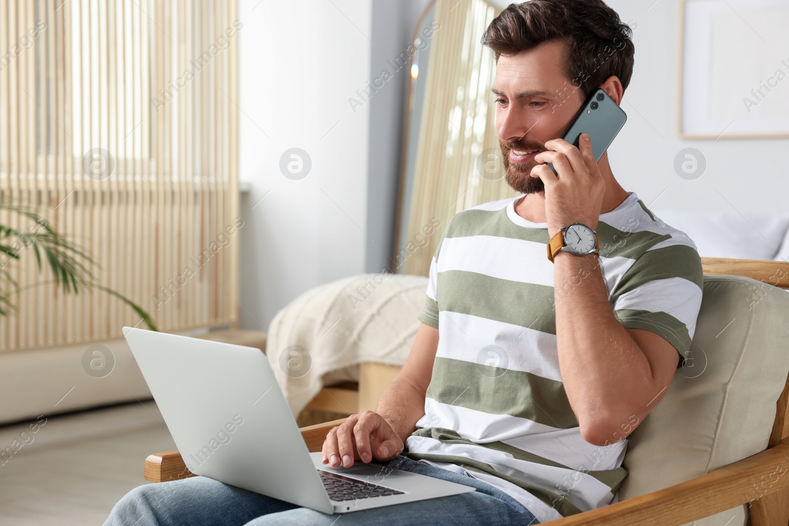 Photo of Handsome man talking on smartphone while working with laptop at home